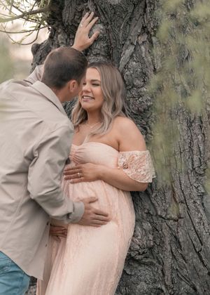Couple sharing a tender moment by a tree, pregnant woman in pink lace off-shoulder gown smiling warmly.