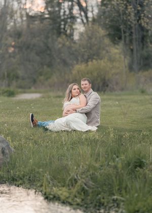 Couple sitting together in a lush green field at sunset, woman in white dress cradled by partner.