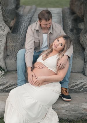 Pregnant couple posing on stone steps, man embracing woman in white maternity dress during outdoor photo shoot.