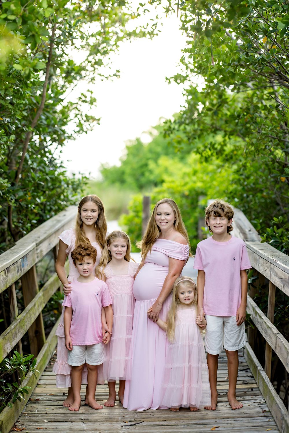 Pregnant woman in pink dress poses with five children on wooden boardwalk surrounded by lush green trees.