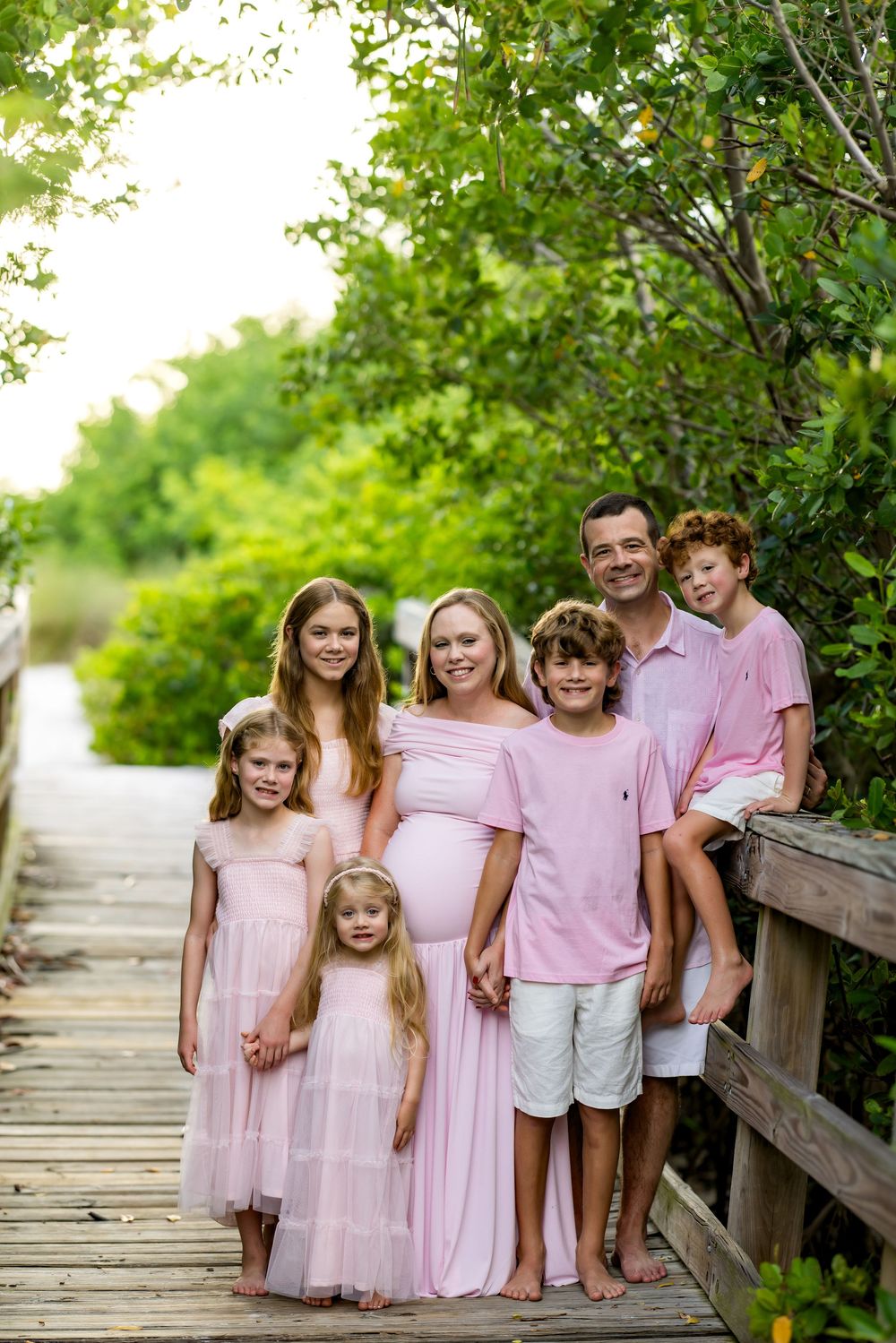 A pregnant woman in pink poses with her family of six on a wooden boardwalk surrounded by lush green trees.