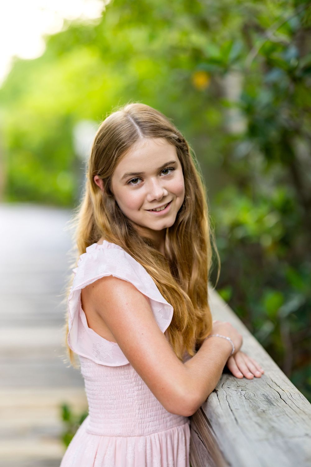 A smiling young girl with long blonde hair in a pink dress leans on a wooden railing outdoors.