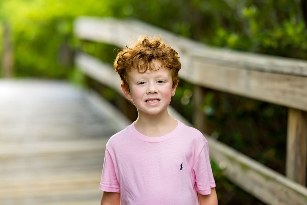 Smiling redhead boy in pink Polo shirt standing on wooden boardwalk surrounded by lush green foliage.