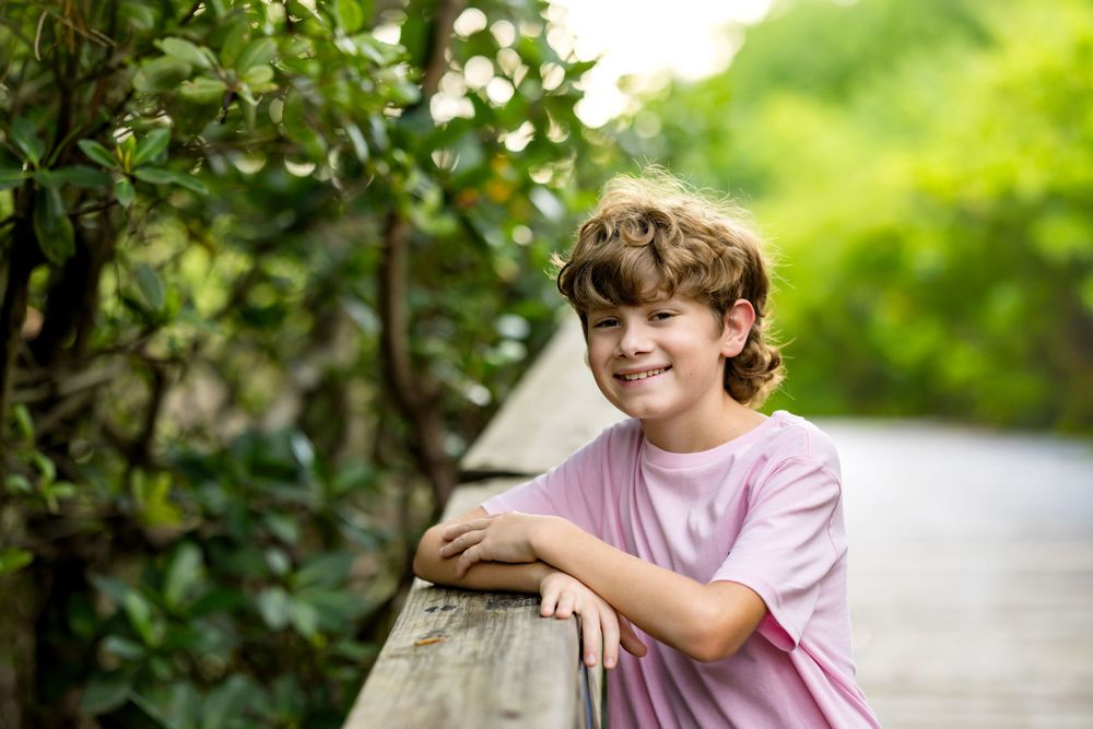 Smiling boy with curly hair leaning on wooden railing surrounded by lush green foliage outdoors.
