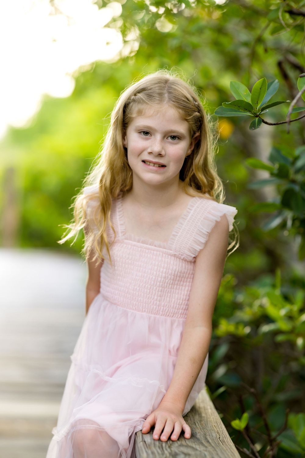 Young girl in pink dress sitting outdoors near green foliage on a sunny day.