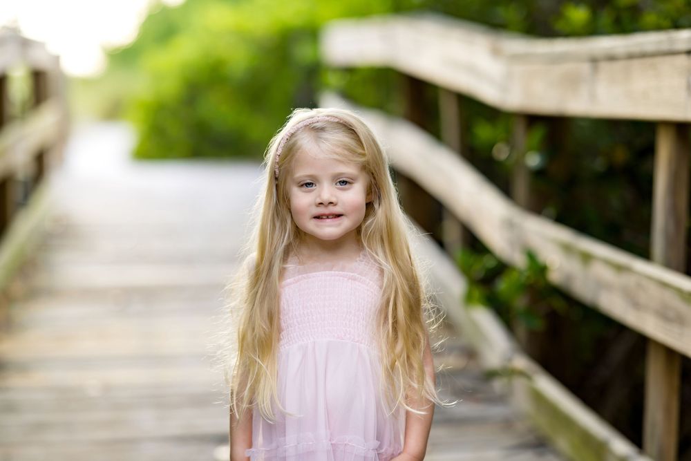 A smiling young girl with long blonde hair wearing a pink dress stands on a wooden boardwalk surrounded by greenery.