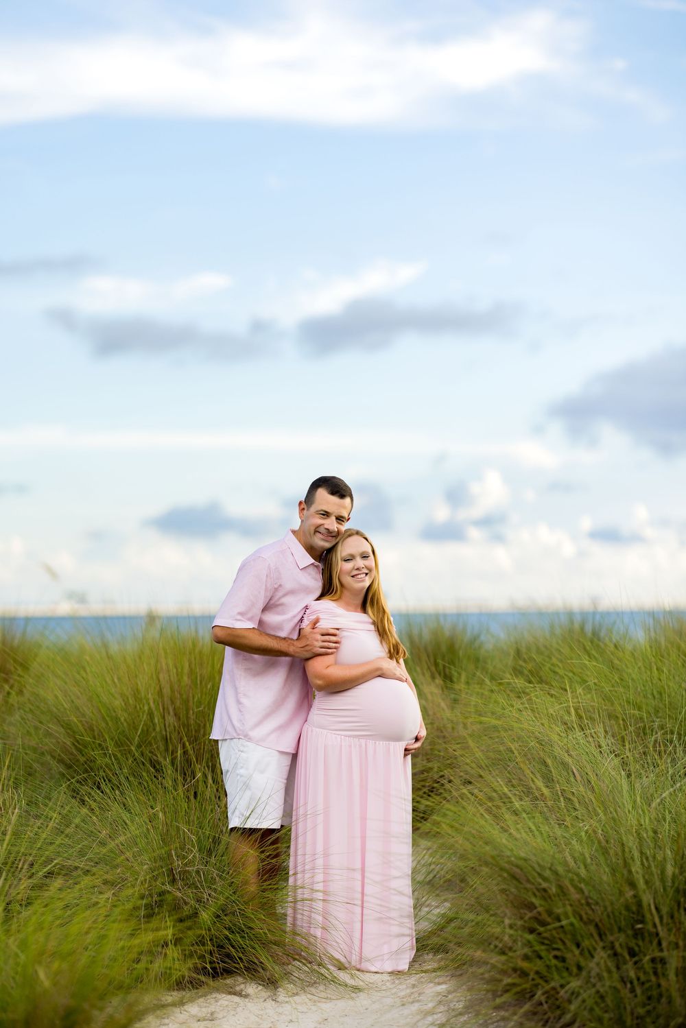 Couple poses on sandy beach path, pregnant woman in pink maxi dress, partner in pink shirt, ocean backdrop.