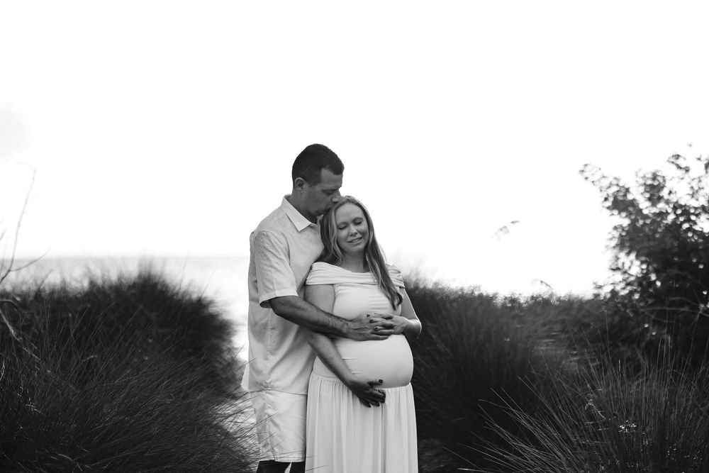 Black and white photo of a pregnant couple embracing outdoors near beach dunes, woman in white dress smiling joyfully.