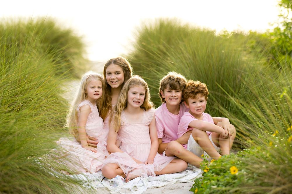 Five children in pink outfits sitting together on a blanket surrounded by tall green grass in a sunny outdoor setting.