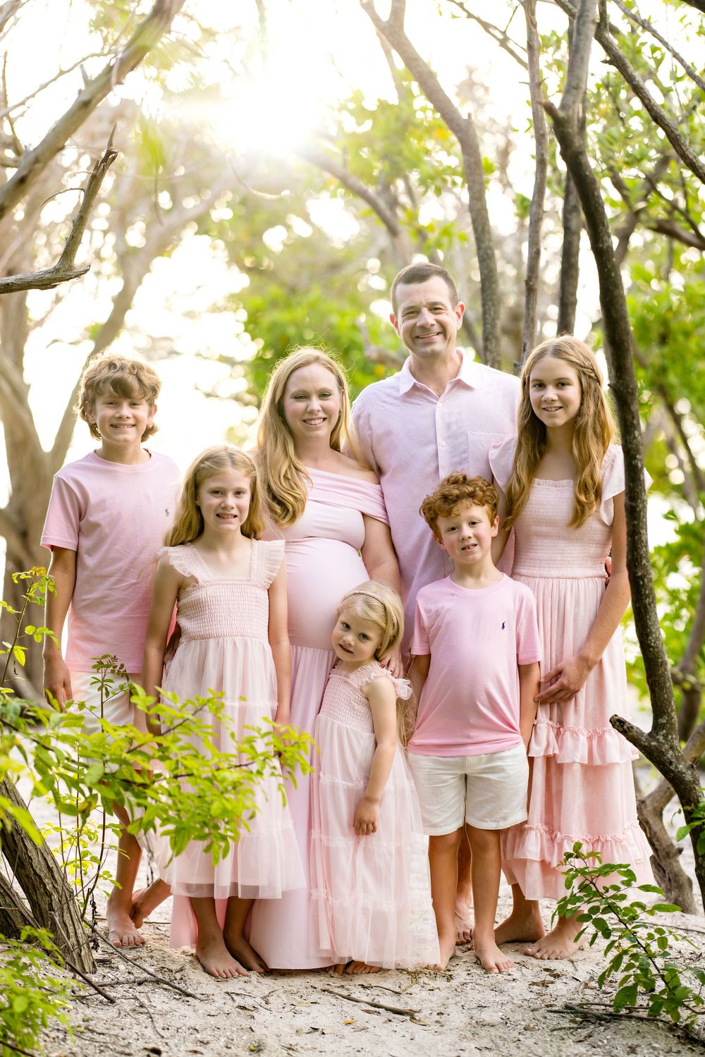 Family of seven in matching pink outfits posing outdoors among trees in a bright, sunlit nature setting.