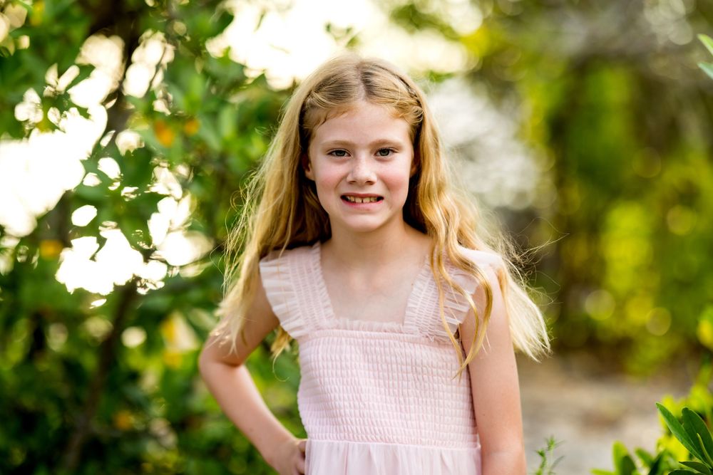 Smiling young girl with long blonde hair wearing a pink dress outdoors among green trees.