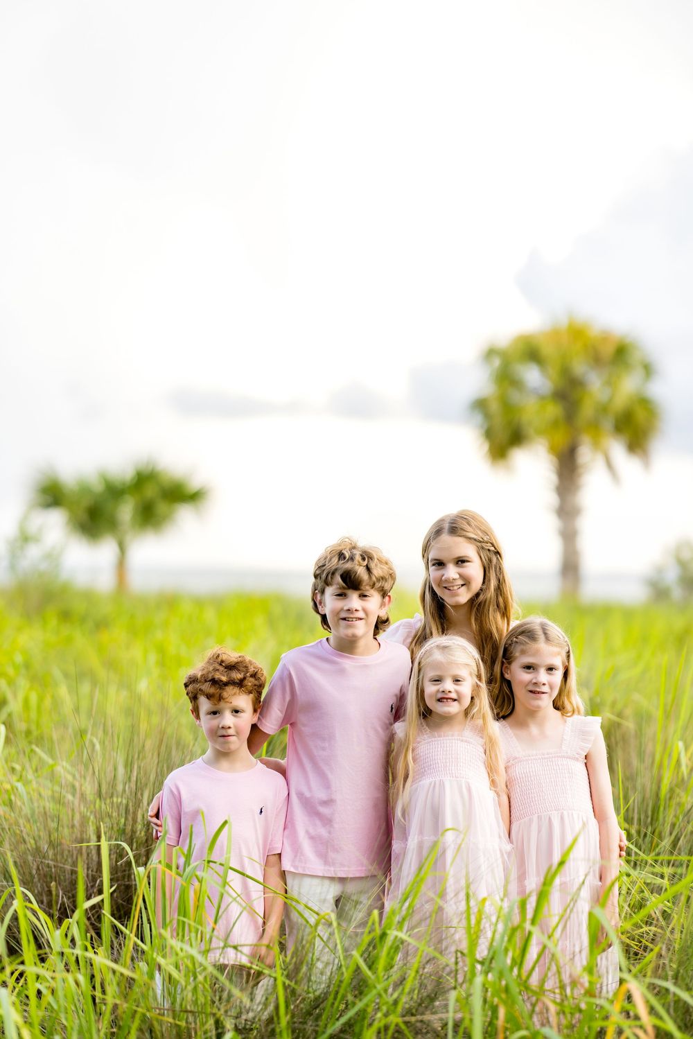 Five children in pink outfits pose together in a lush green field with palm trees in the background.