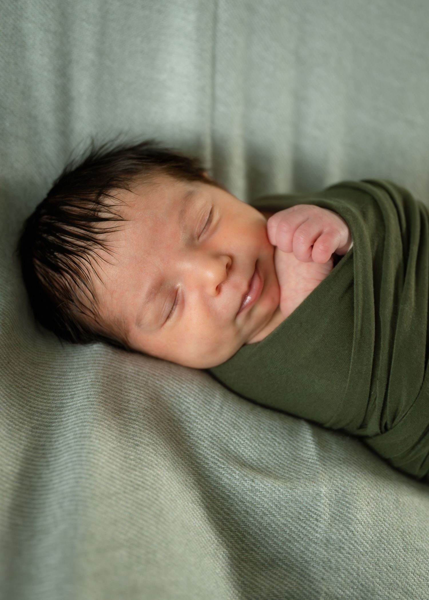 Peaceful newborn baby swaddled in olive green wrap, sleeping soundly with dark hair and tiny fist visible.