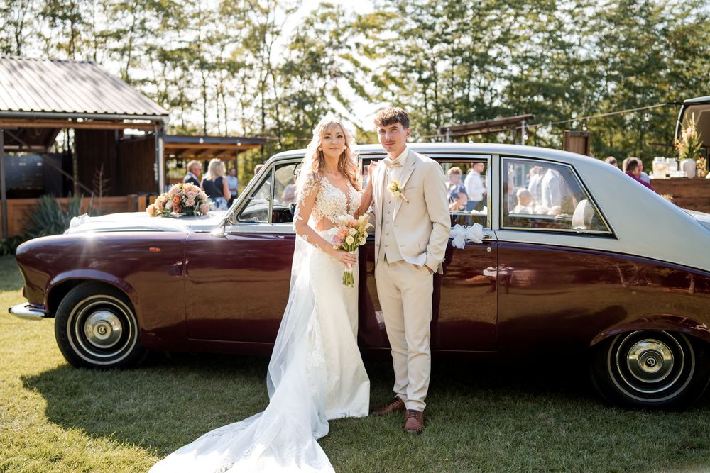 Bride and groom posing beside a vintage maroon Rolls-Royce at an outdoor wedding venue.