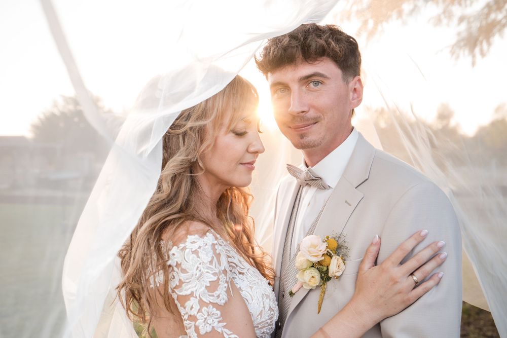 Bride and groom share romantic moment under flowing veil with golden sunset backlight at outdoor wedding.