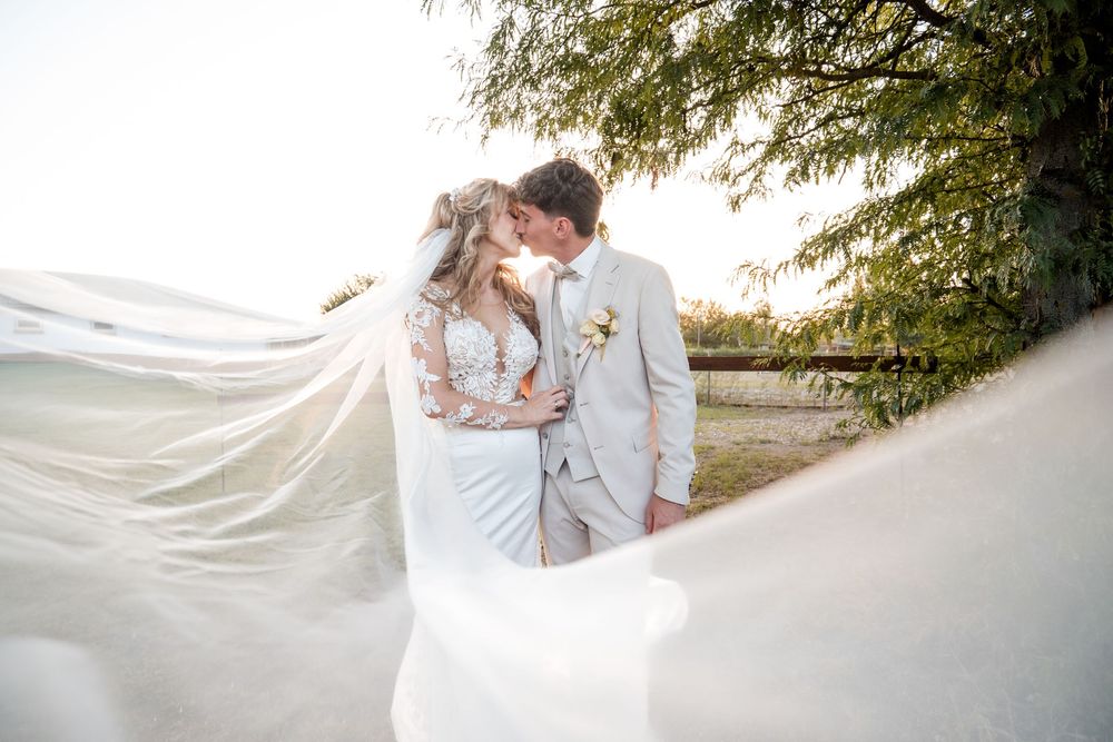 Bride and groom sharing a romantic kiss outdoors with flowing white veil billowing in the wind on their wedding day.