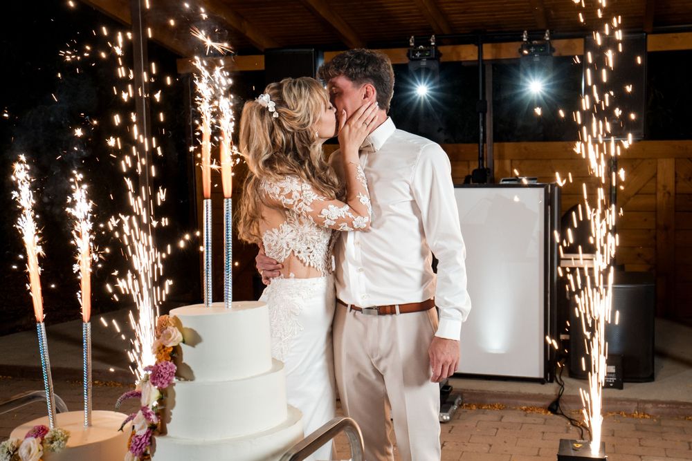 Newlyweds share a kiss beside their wedding cake surrounded by sparkling fountain fireworks at their reception.