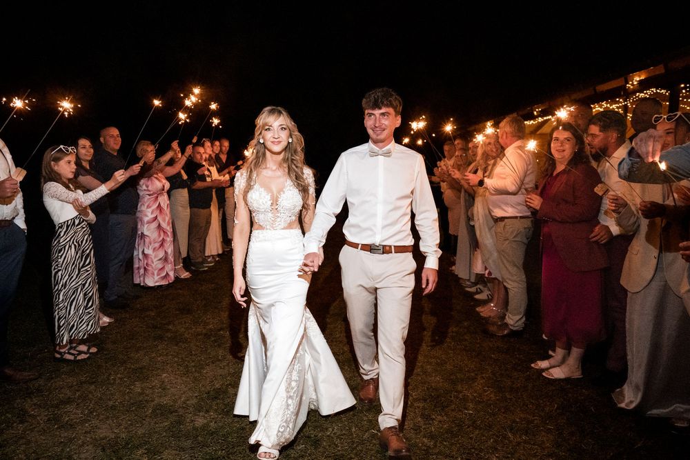 Newlywed couple walks through sparkler exit at outdoor night wedding celebration surrounded by cheering guests.