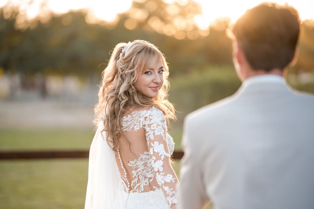 Bride in lace wedding gown glances over shoulder at sunset, golden light illuminating her elegant hairstyle.