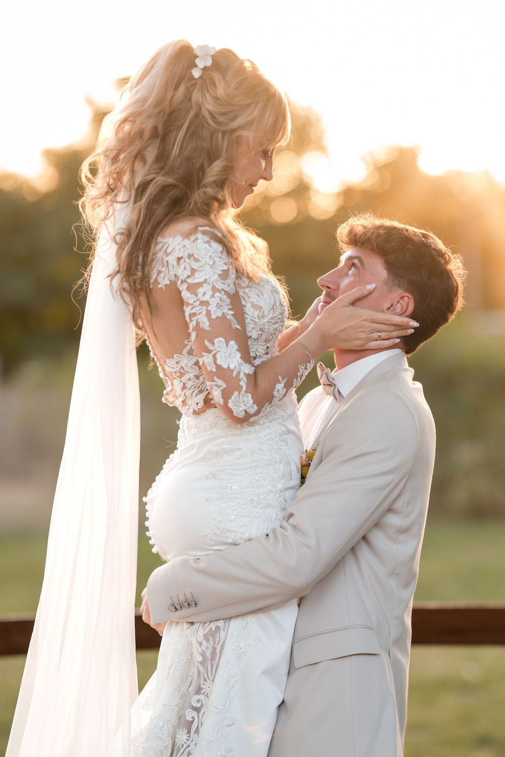 Groom lifts bride in lace wedding dress at golden hour, sharing a romantic moment outdoors.