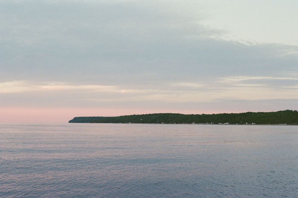 Serene coastal landscape at dusk with calm blue waters and a tree-lined peninsula under a soft pink and gray sky.