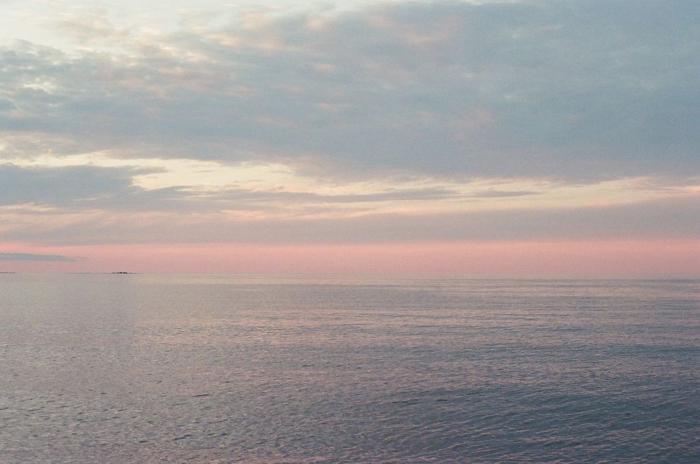 Serene ocean sunset with pastel pink and blue sky reflecting on calm water, with a distant ship on the horizon.