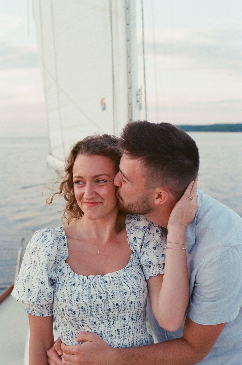 A couple shares a romantic moment on a sailboat at sunset, man kissing woman's cheek on calm ocean waters.