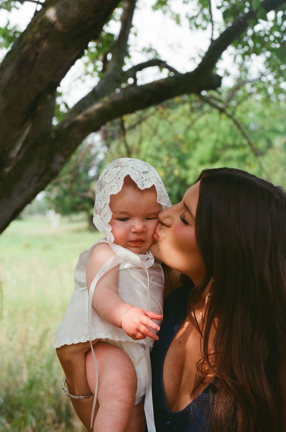 A mother kissing her baby wearing a white lace bonnet outdoors under a tree.