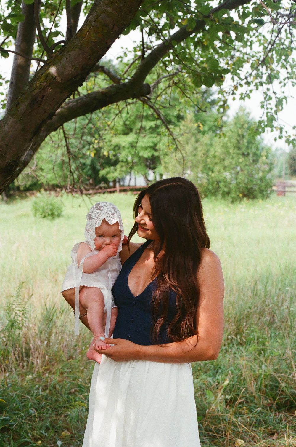 A mother in a navy top and white skirt holds her baby in a lace bonnet outdoors near a tree.