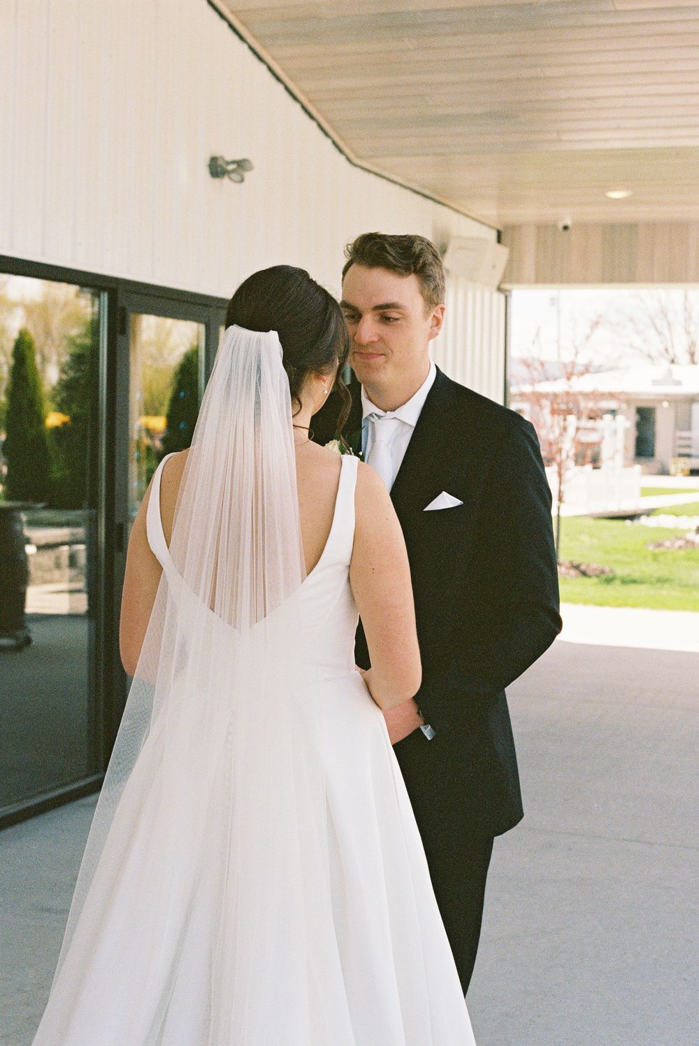 Bride in white gown and veil shares intimate moment with groom in black tuxedo outside wedding venue.