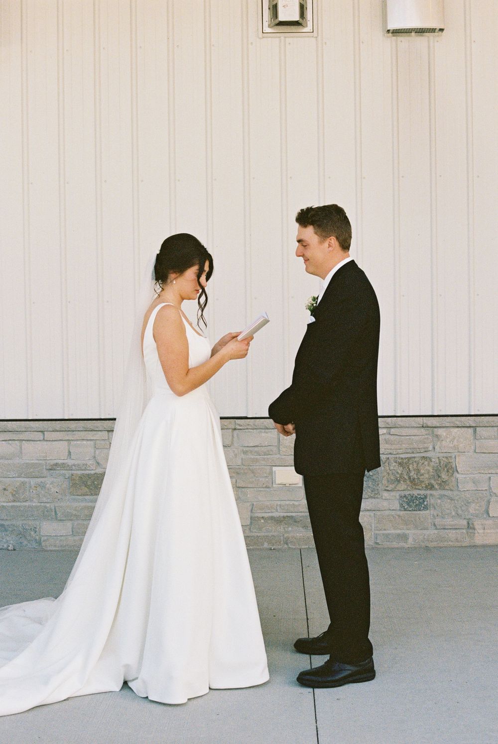 Bride in elegant white gown reads vows to groom in black suit during outdoor wedding ceremony.