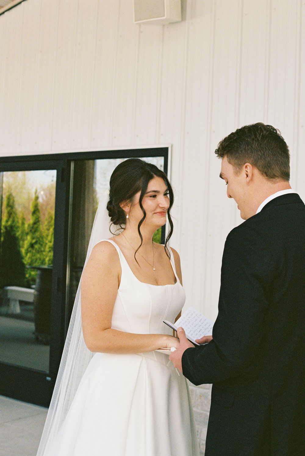 Bride smiles warmly at groom as they exchange personal wedding vows outside a white barn venue.