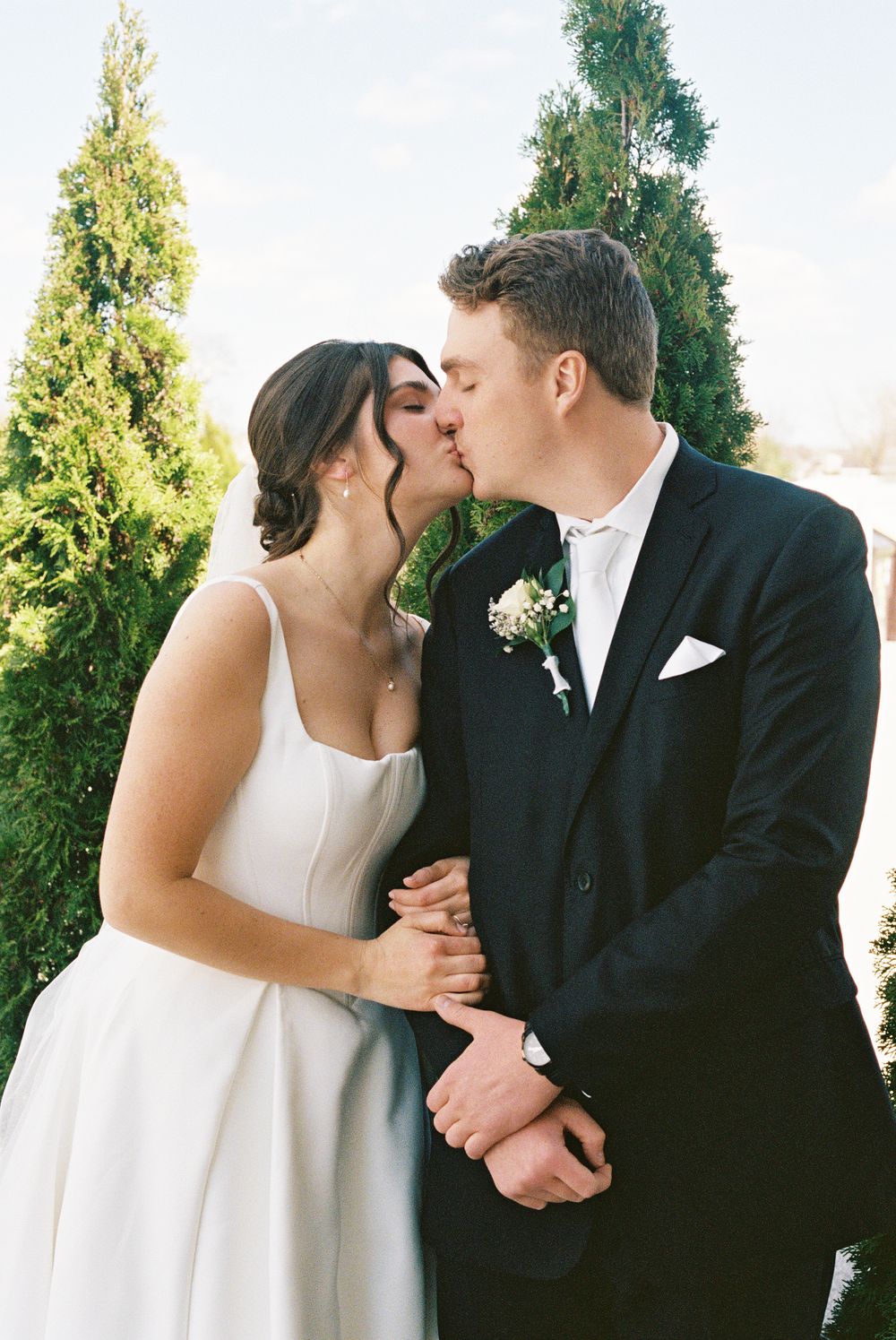 A bride and groom share a romantic kiss outdoors, dressed in a white wedding gown and black tuxedo.