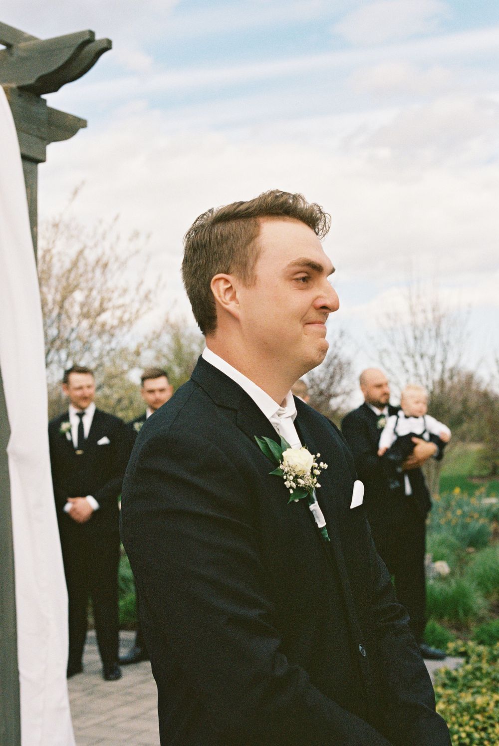 Emotional groom in black tuxedo with boutonniere watches bride walk down the aisle at outdoor ceremony.