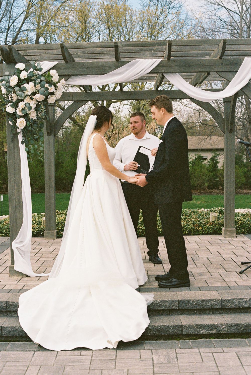 Bride and groom exchange vows under floral pergola with white draping and rose arrangements at outdoor ceremony.