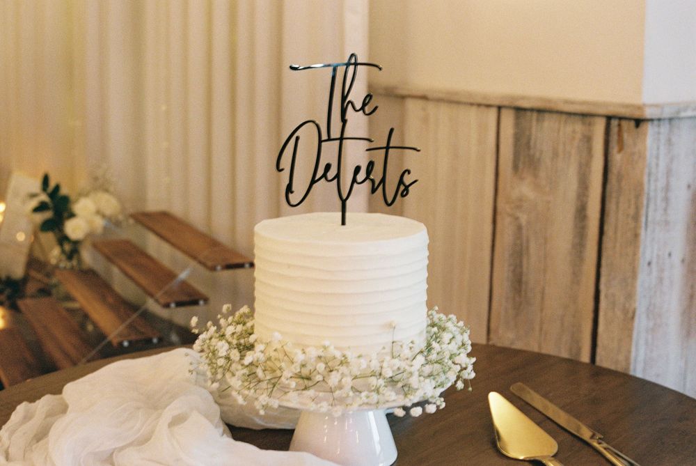 Elegant white wedding cake topped with The Deterts cake topper, decorated with baby's breath flowers.