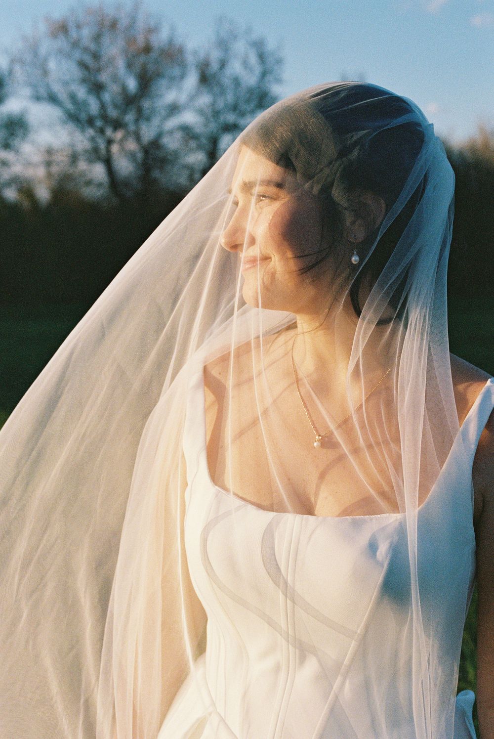 A bride in a white gown smiles softly under a flowing veil in golden hour light outdoors.