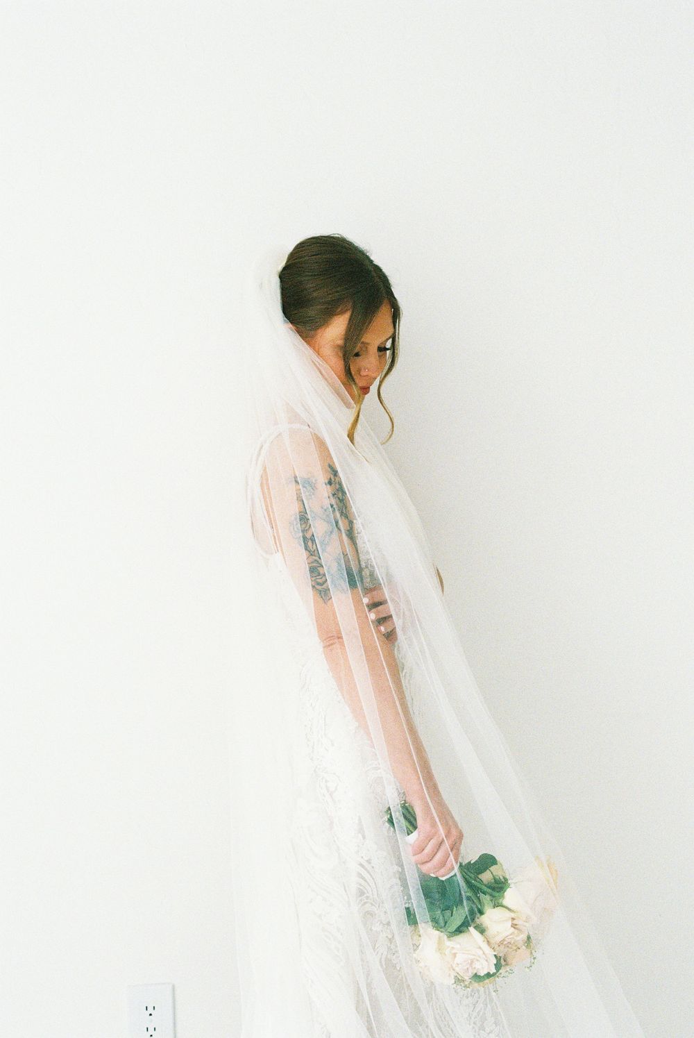 Bride in white lace gown and flowing veil holds bouquet of roses, gazing down against a white wall.
