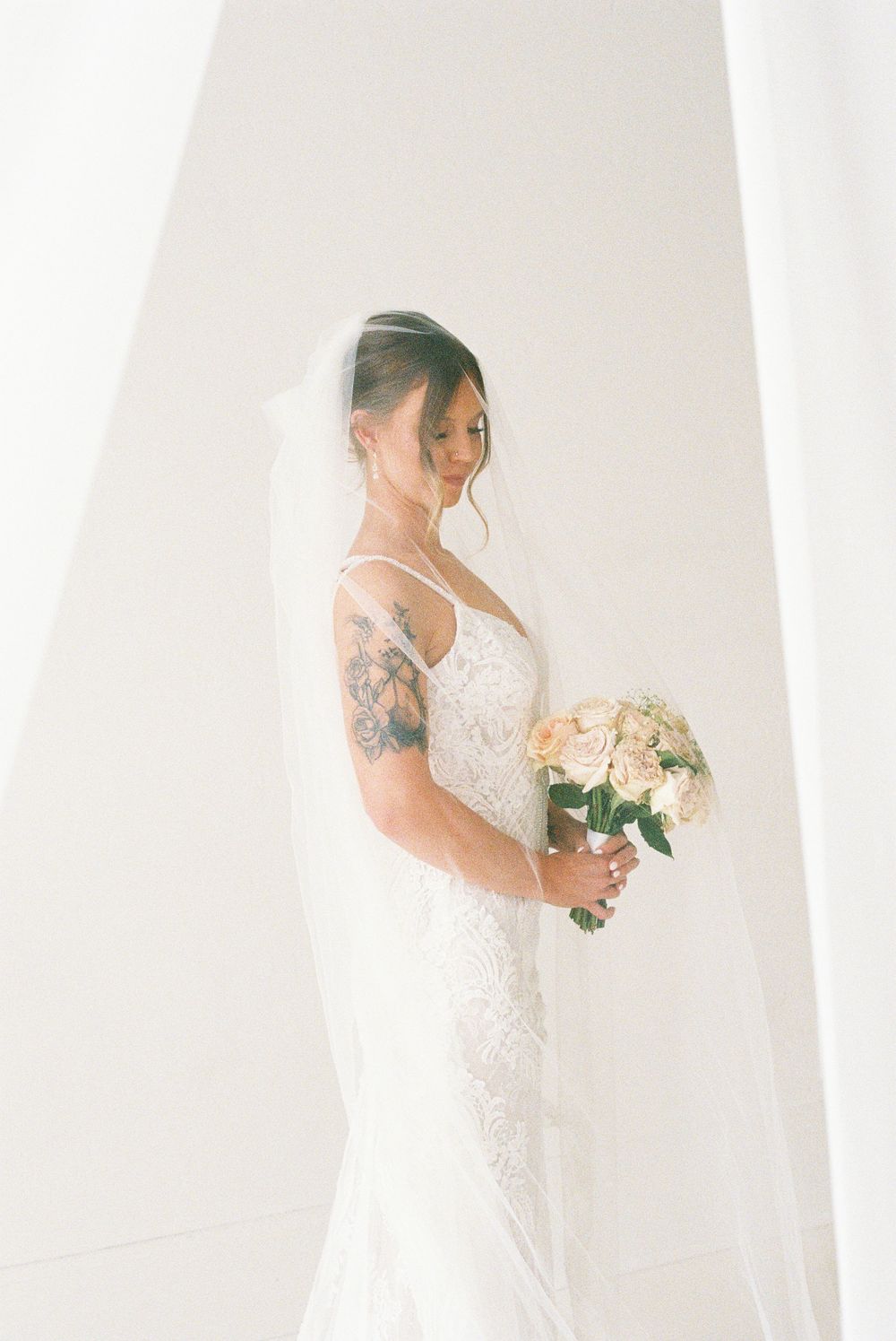 Bride in lace gown and veil holds blush bouquet, tattoo visible on arm, standing in soft white light.