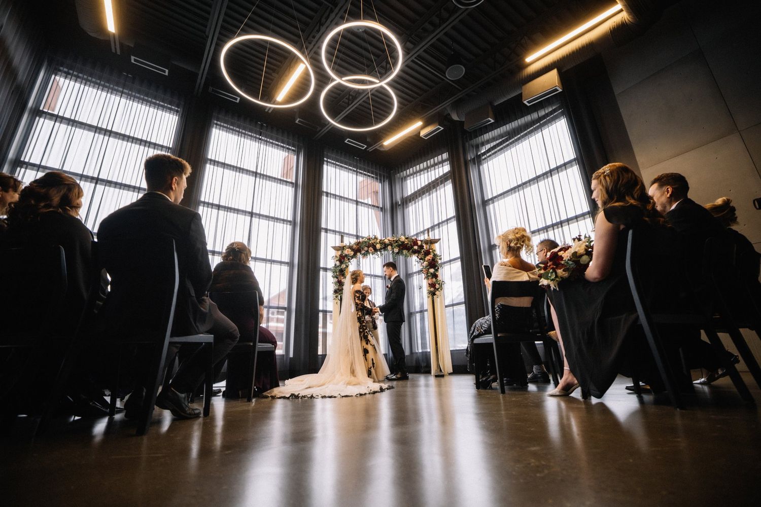 Low wide view of a wedding ceremony at Mainspace in Canmore under circular ring lights and tall windows