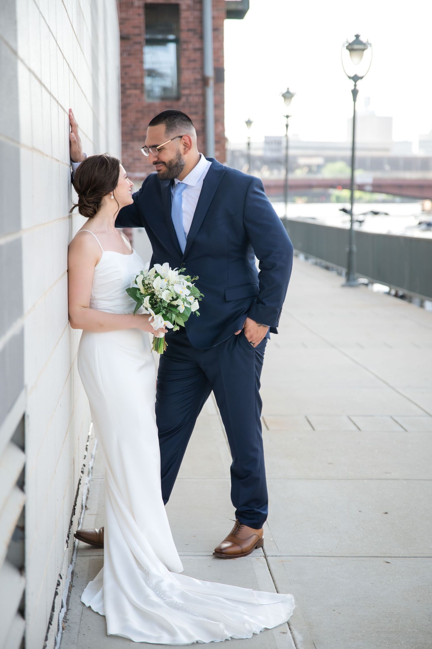Bride in a white satin gown holding a white bouquet leans against a wall beside a groom in a navy suit on a riverside walkway.