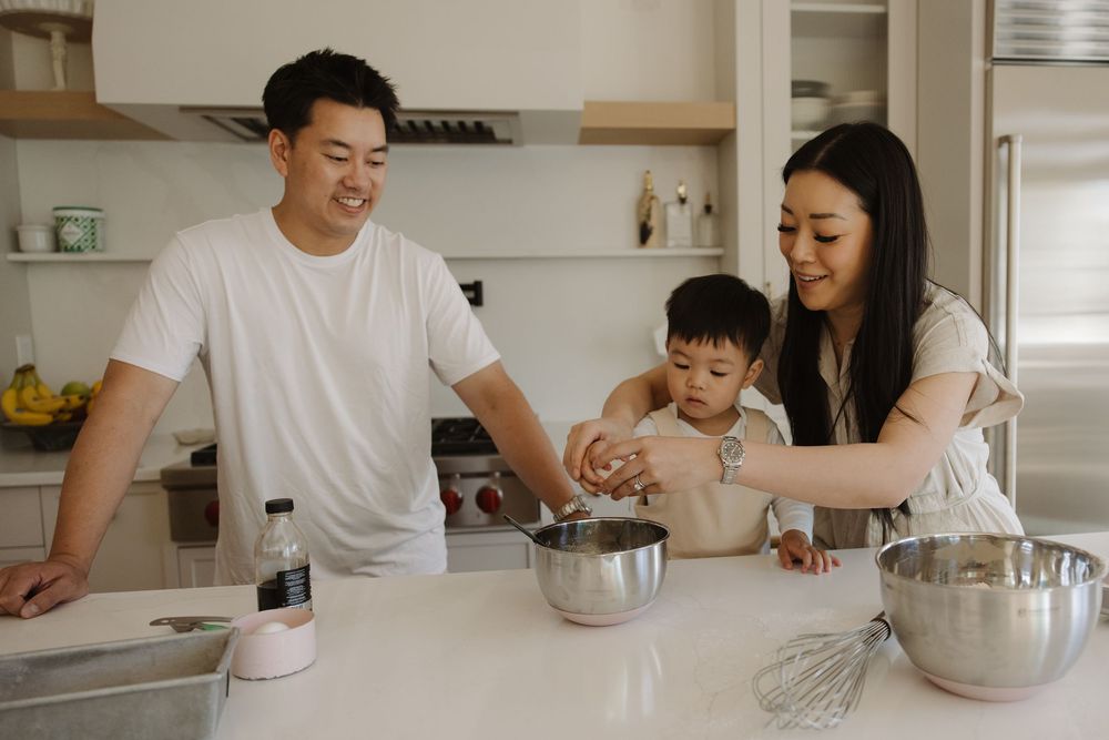 Family cooking together in a bright kitchen, parents helping toddler crack eggs into a mixing bowl.