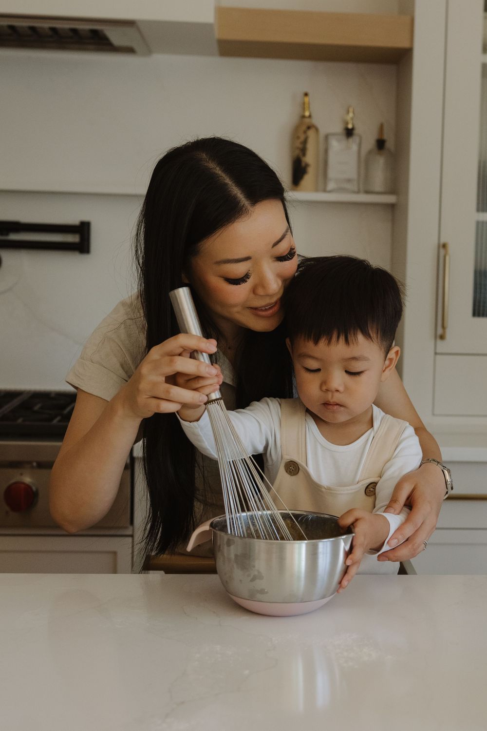 A mother and toddler cooking together in a kitchen, whisking ingredients in a metal bowl.