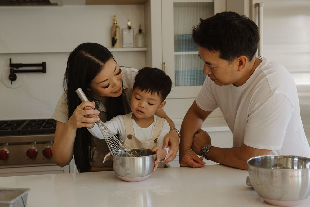 Asian family cooking together in a modern white kitchen, parents teaching toddler to whisk ingredients in a mixing bowl.