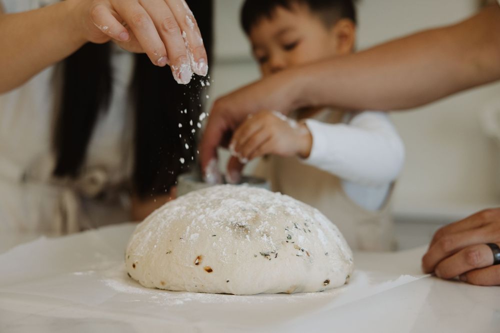 Hands dusting flour on fresh bread dough on a white surface with a child helping in the background.