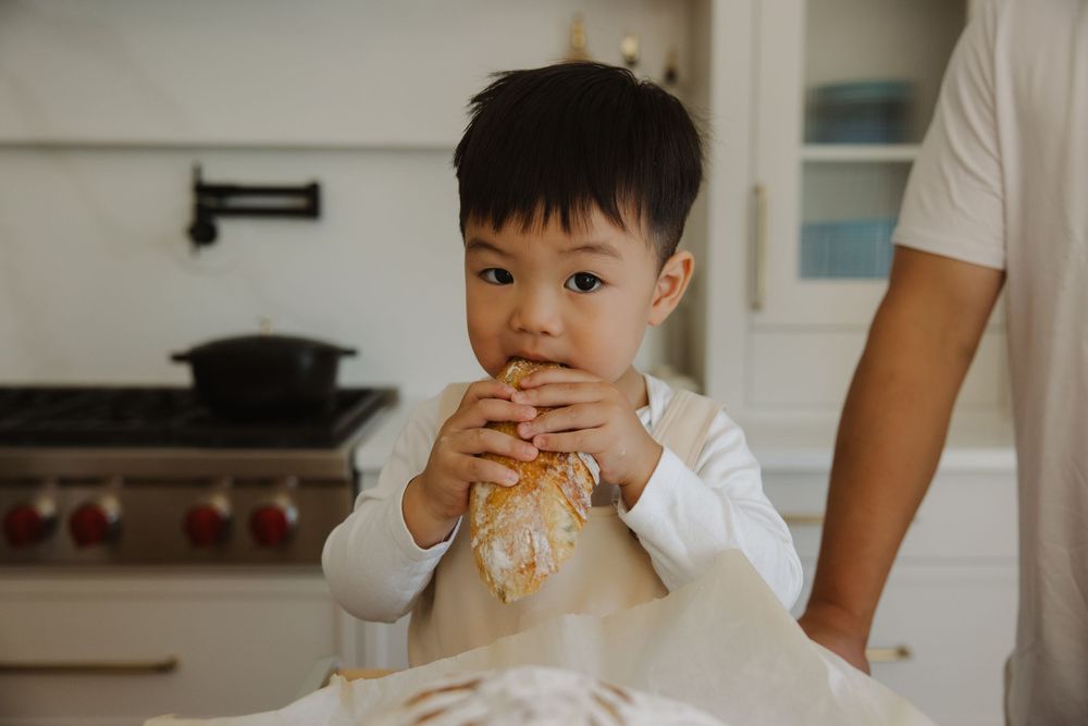 Cute toddler boy eating bread dough in a modern white kitchen while baking with an adult.
