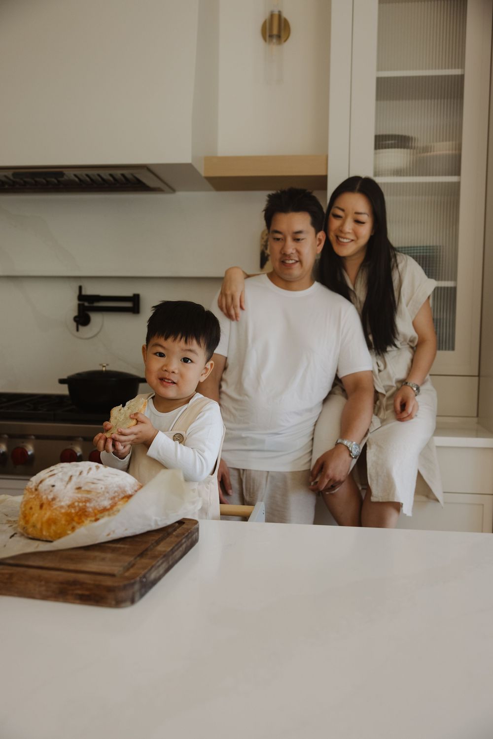 Happy family in a modern white kitchen, parents smiling as young son enjoys fresh baked bread they made for in home family pictures