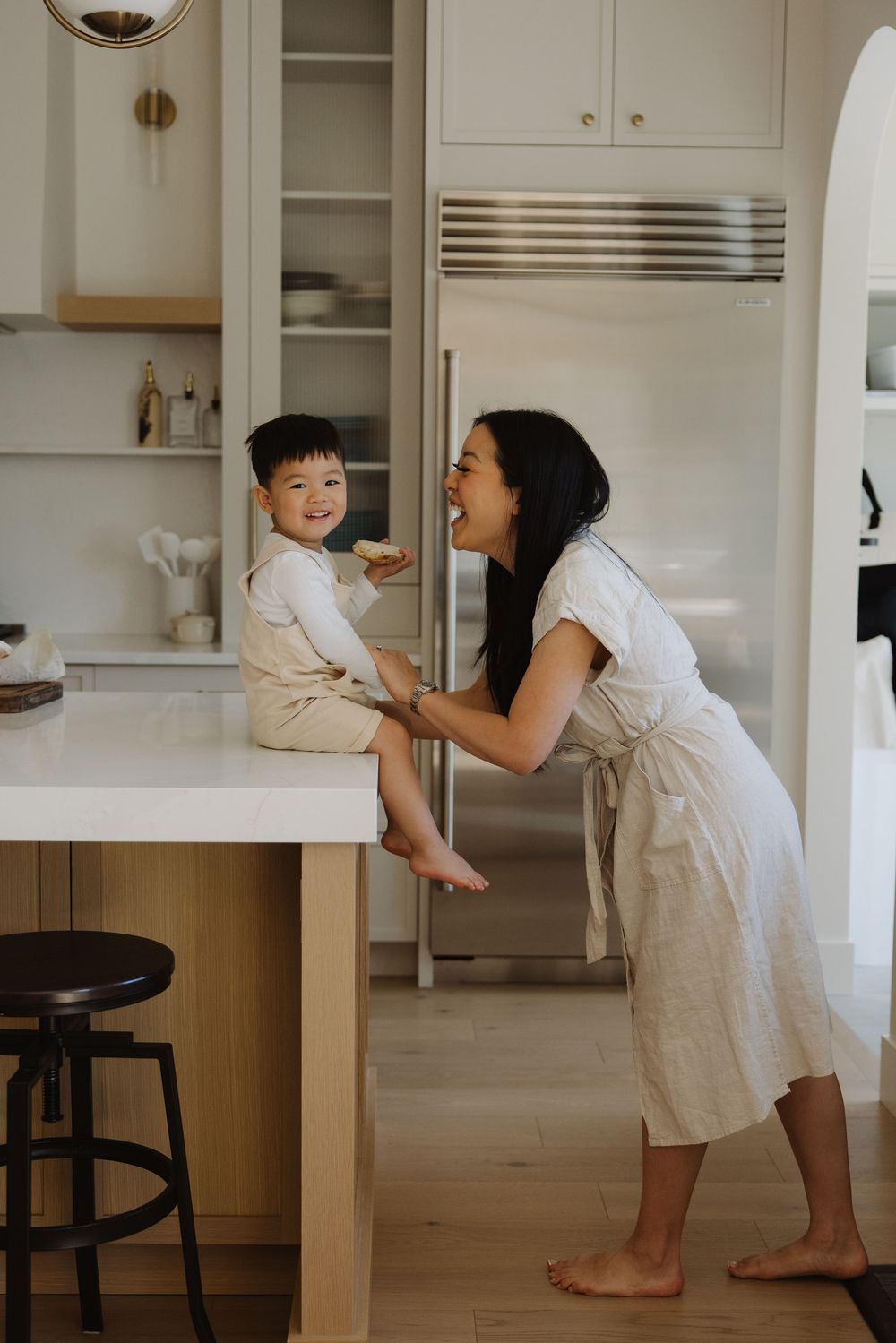 A mother laughs with her toddler son sitting on a kitchen counter in a bright, modern white kitchen during thier calgary in home photoshoot