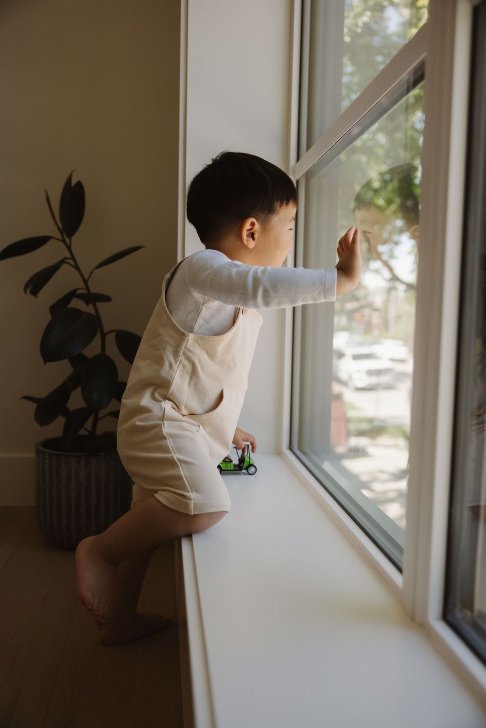 Toddler in beige overalls standing on tiptoes, looking out a bright window while holding a toy car.