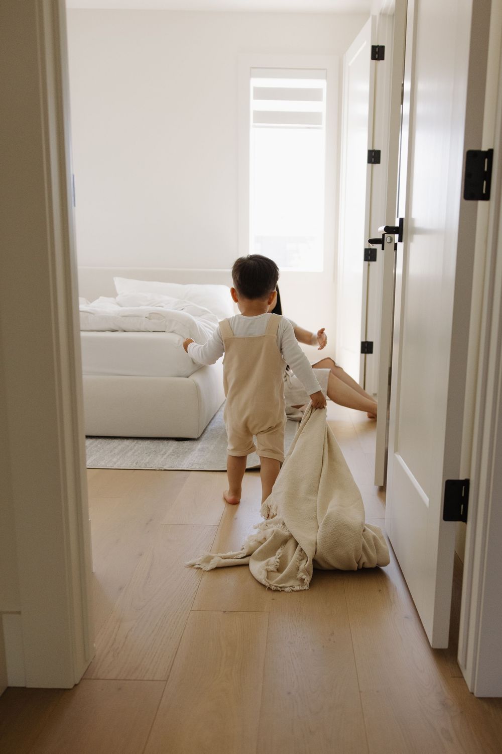 Toddler in overalls dragging a blanket through a bright white bedroom doorway to snuggle mom during in home family pictures in yyc